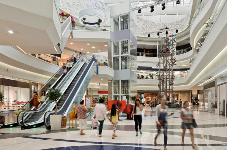 Shoppers walking through large. bright, 3-story mall atrium, with elevator and escalator.
