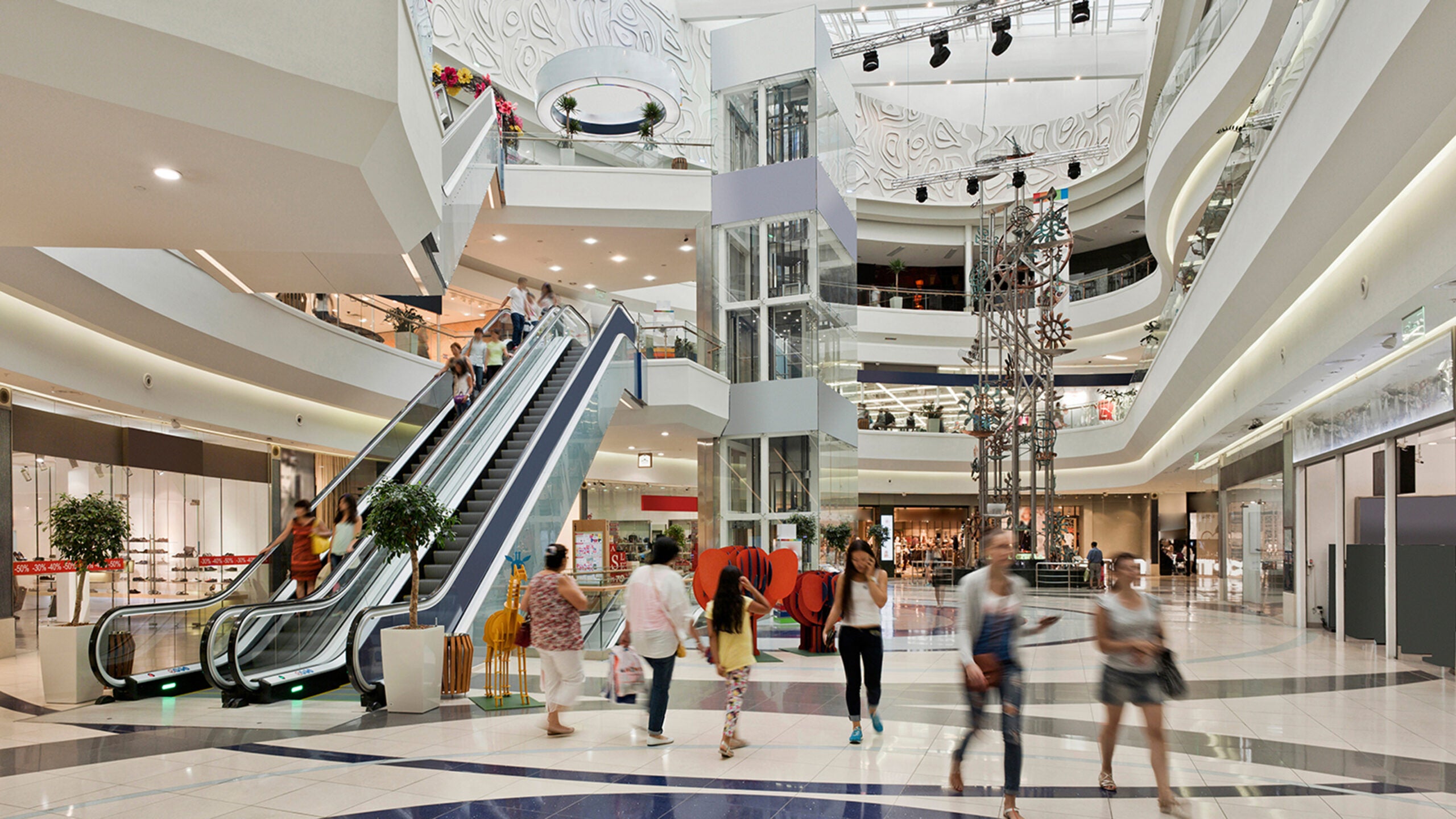 Shoppers walking through large. bright, 3-story mall atrium, with elevator and escalator.